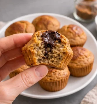 Mano sosteniendo magdalena casera con interior esponjoso y chocolate fundido, con más magdalenas al fondo